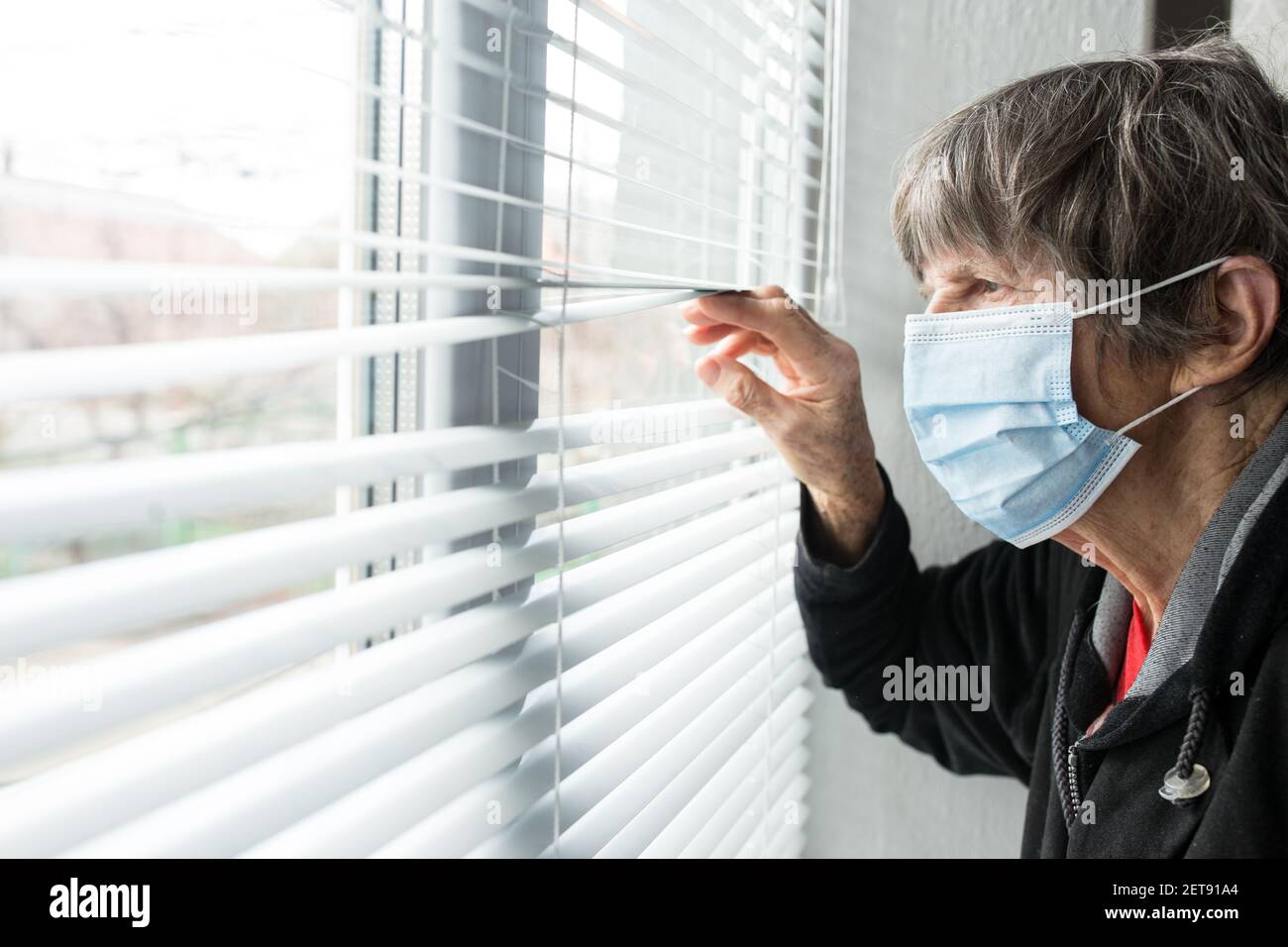 Una donna anziana in una maschera medica guarda fuori la finestra. Foto Stock