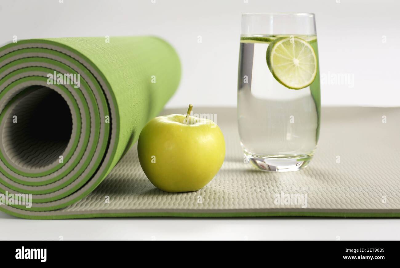 Yoga fitness, stile di vita. Yoga laminato verde mat, mela verde, bicchiere d'acqua con limone su sfondo bianco. Posiziona per il testo. Stile di vita sano, fitness, concetto sportivo Foto Stock