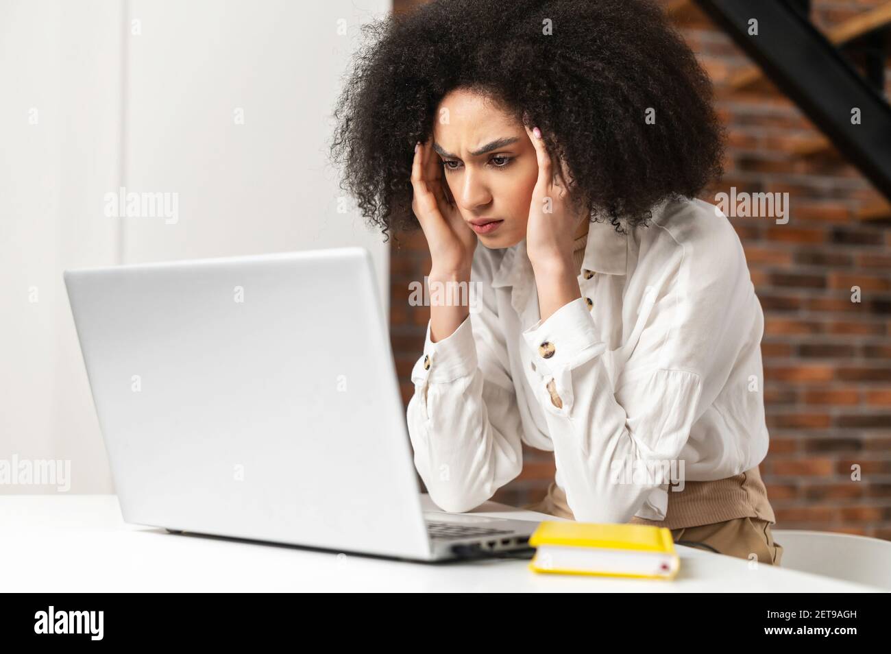 Giovane donna disorientata che lavora in ufficio è sotto stress emotivo, guardando il laptop, tenendo le mani ai suoi templi, strizzando la fronte, overworked, il computer è rotto, che cosa è successo Foto Stock