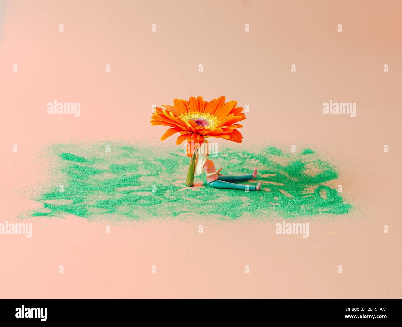 Una ragazza bionda seduta su una sabbia verde all'ombra di un gigantesco fiore di gerbera. Concetto creativo di primavera o estate. Foto Stock