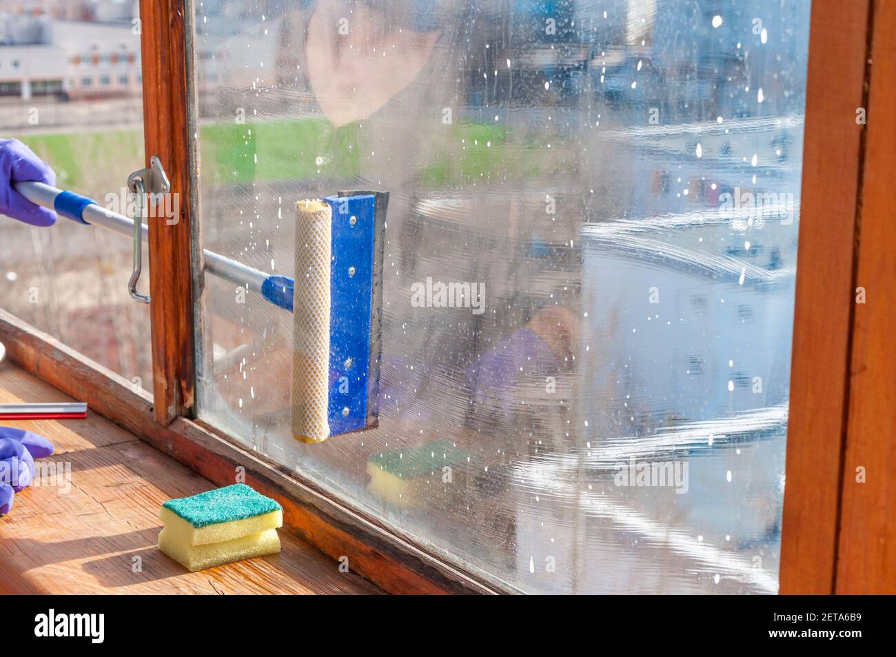 Una giovane donna irriconoscibile in guanti blu lava il balcone finestra dall'esterno con uno speciale raschiatore a manico lungo Foto Stock