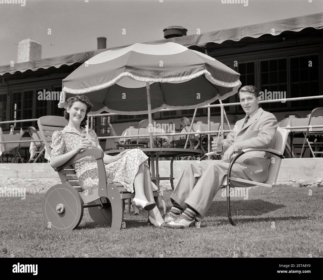 1940 COPPIA UOMO E DONNA MOGLIE MARITO SEDUTA IN PRATO SEDIE BERE BEVANDE  ANALCOLICHE SOTTO UN OMBRELLO SORRIDENTE GUARDANDO FOTOCAMERA - F5707  HAR001 HARS VECCHIO STILE 1 GIOVANE VACANZA ADULTA GIOIA SODDISFAZIONE