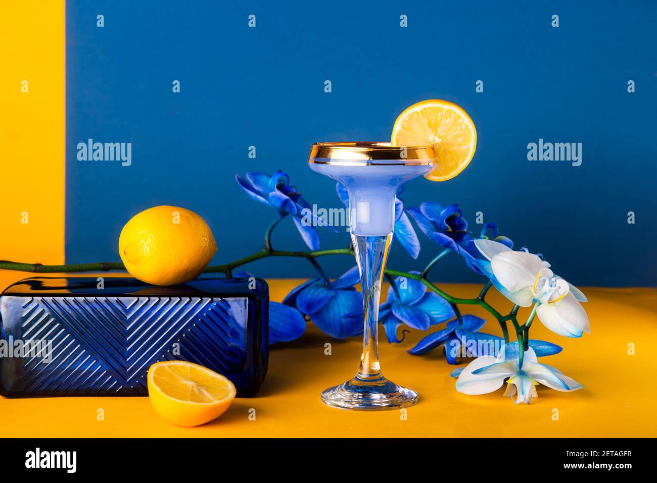 Cocktail tropicale in vetro con fresco decor al limone, fiori di orchidea blu, podio a trama di vetro su sfondo giallo e blu. Minimalismo alla moda geometrico Foto Stock