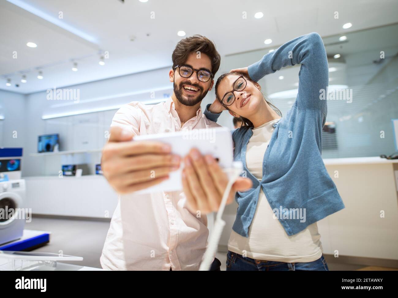 Giovane coppia allegra sta mettendo in posa per un selfie mentre prova la macchina fotografica su un telefono in un negozio di tecnologia luminoso. Foto Stock