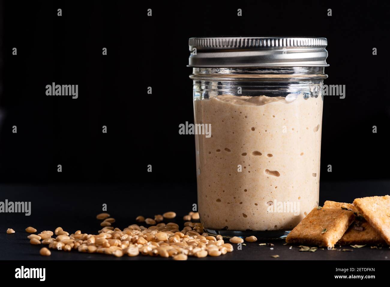 Impasto agitato nel vaso di vetro, bacche di grano duro, cracker fatti in casa su fondo nero. Foto Stock