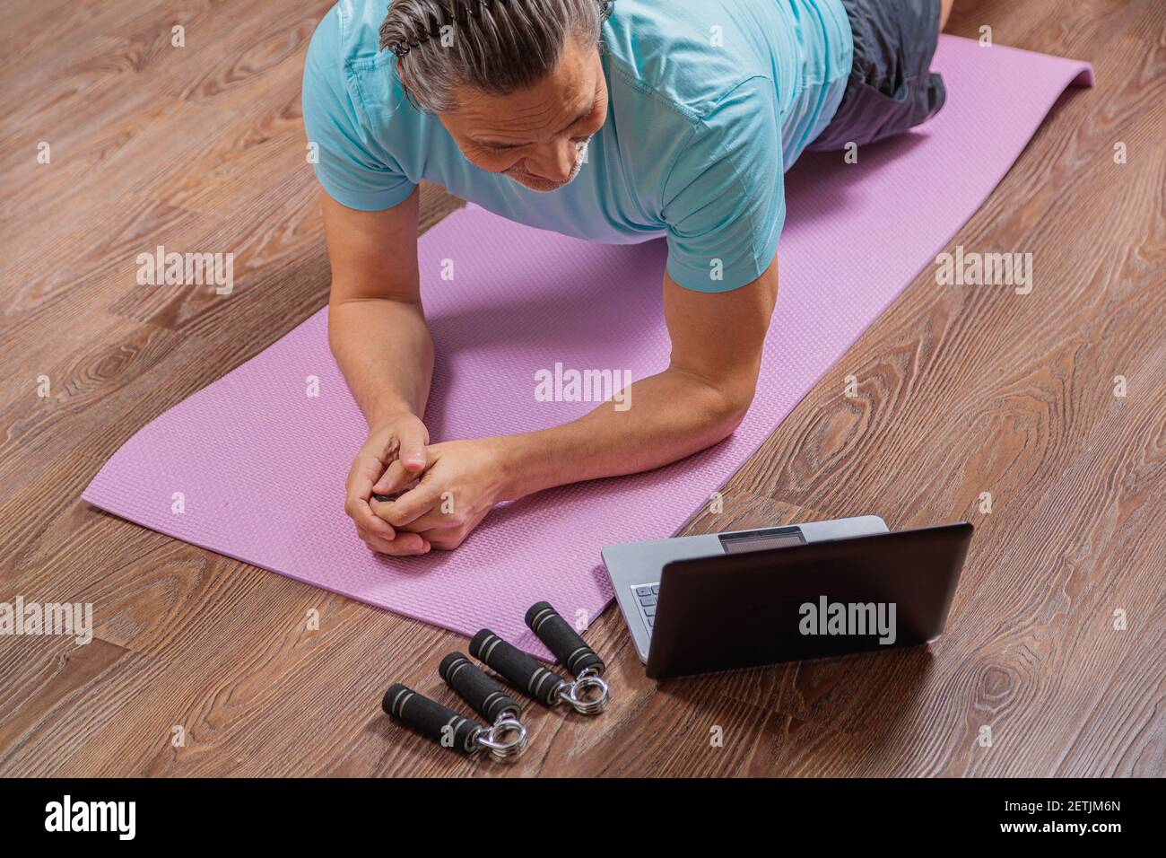 l'uomo di 50 anni esegue esercizi mentre si trova sul tappeto a casa, guardando il computer. Durante una pandemia, una persona si allena in un appartamento tramite il Foto Stock