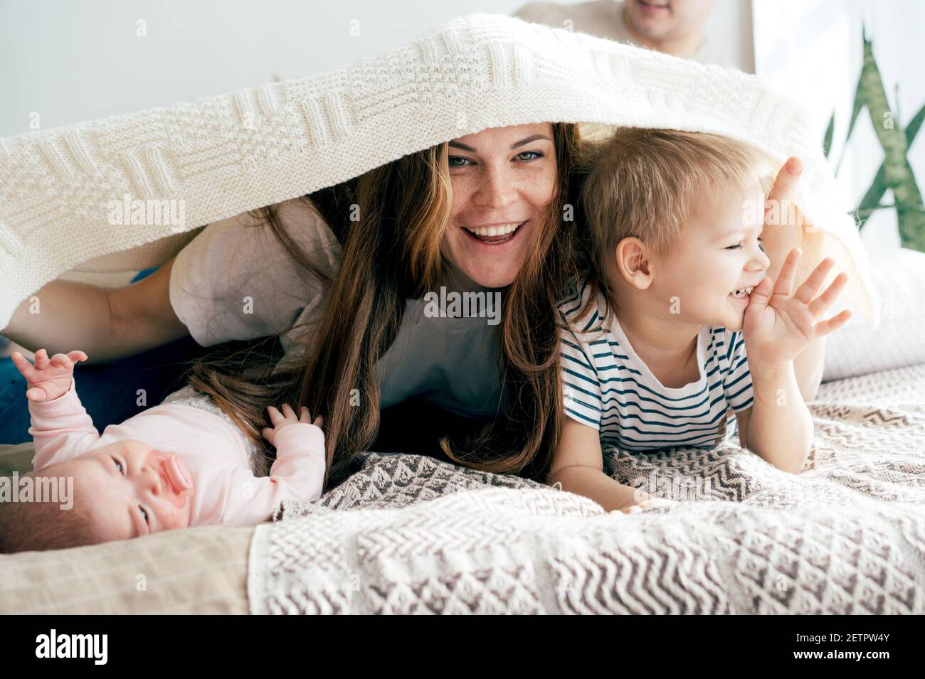 La mummia felice gioca con i bambini sul letto e copre i bambini e se stessa con una coperta. Divertimento per tutta la famiglia. Foto Stock