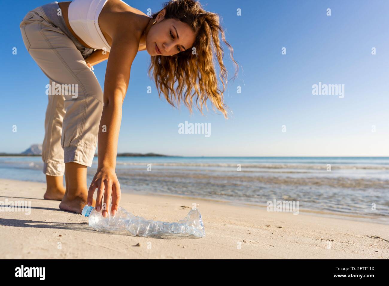 Giovane donna ben istruita e sensibile a questioni ambientali raccoglie una bottiglia di plastica vuota abbandonata in riva al mare per preservare l'oceano da Foto Stock