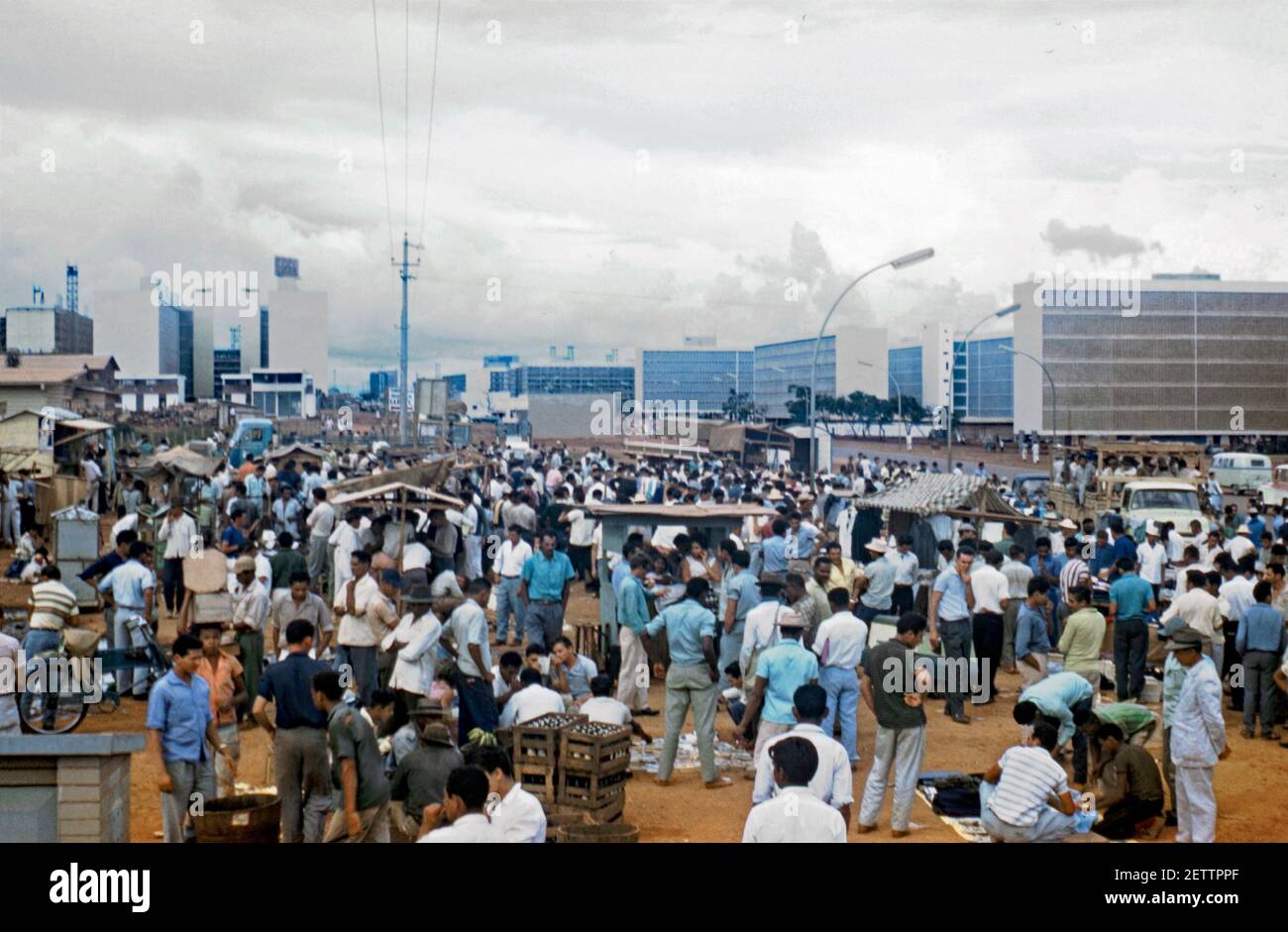 Il vecchio incontra il nuovo - un mercato di strada tradizionale e affollato nella moderna ambientazione di Brasília, Brasile 1961. Le condizioni opache non hanno messo le folle, dominate molto dagli uomini. L'architettura della città moderna incompiuta è vista sullo sfondo. Brasília è la capitale federale del Brasile. La città si trova negli altopiani brasiliani, nella regione centro-occidentale del paese. Brasília fu una città progettata da Lúcio Costa, Oscar Niemeyer e Joaquim Cardozo nel 1956. Questa immagine proviene da una vecchia trasparenza di colore dilettante da 35 mm. Foto Stock