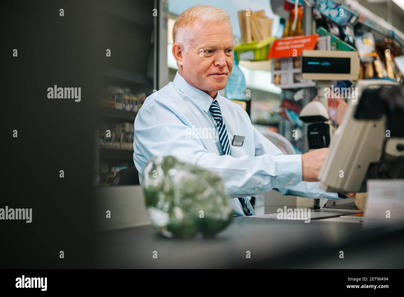 Responsabile del negozio di alimentari seduto al banco del check-out e guardando qualcosa sul computer. Uomo anziano che lavora al banco di cassa del supermercato. Foto Stock