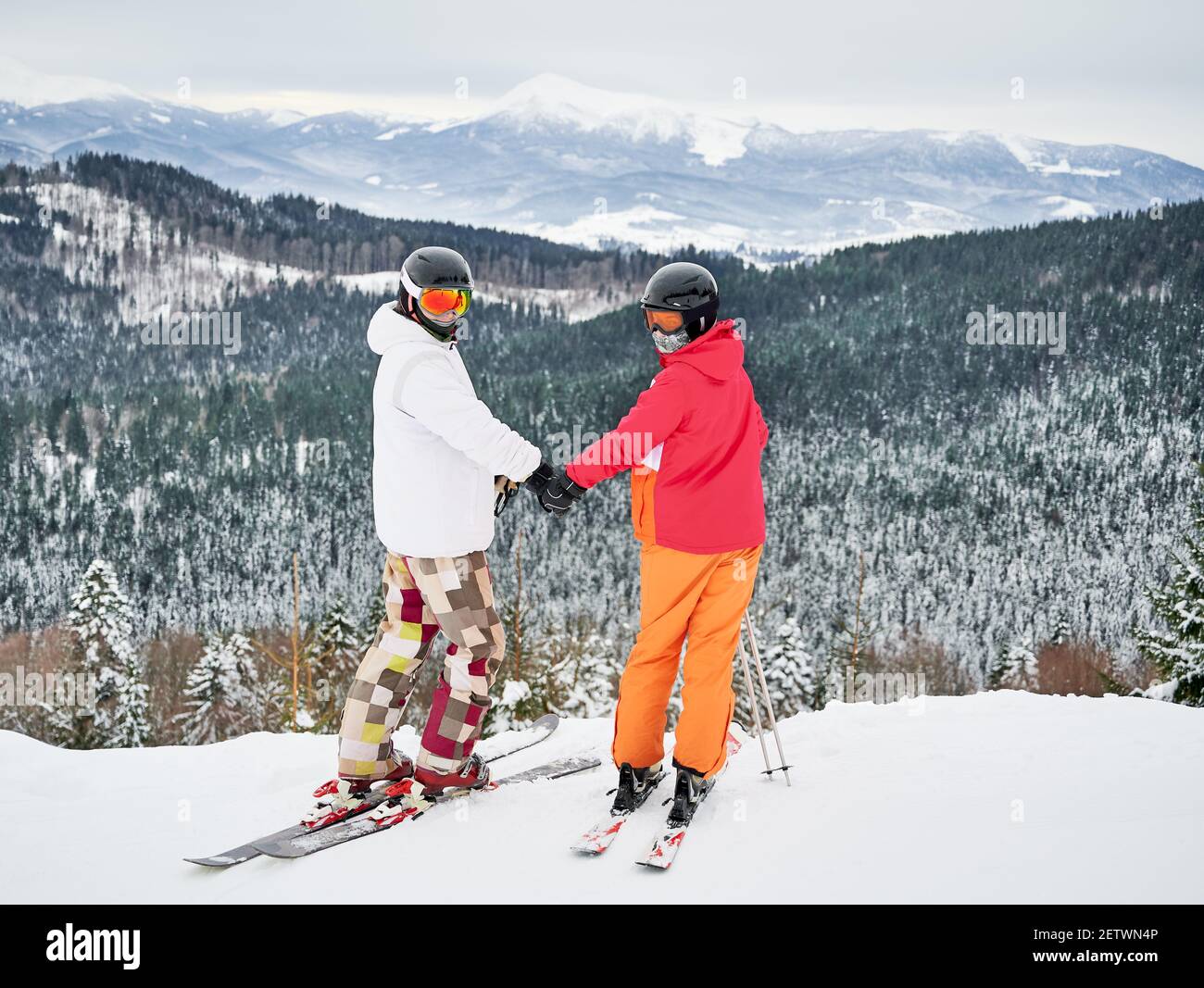 Sciatori uomo e donna che tengono le mani e guardano la macchina fotografica mentre si levano in piedi sulla collina innevata. Giovane coppia sciare insieme durante la giornata invernale in montagna. Concetto di attività sportive invernali, relazioni Foto Stock