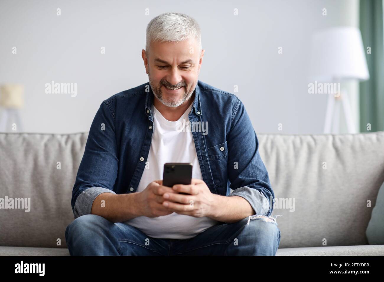 Sorridente uomo dai capelli grigi maturo che usa il telefono cellulare a casa Foto Stock