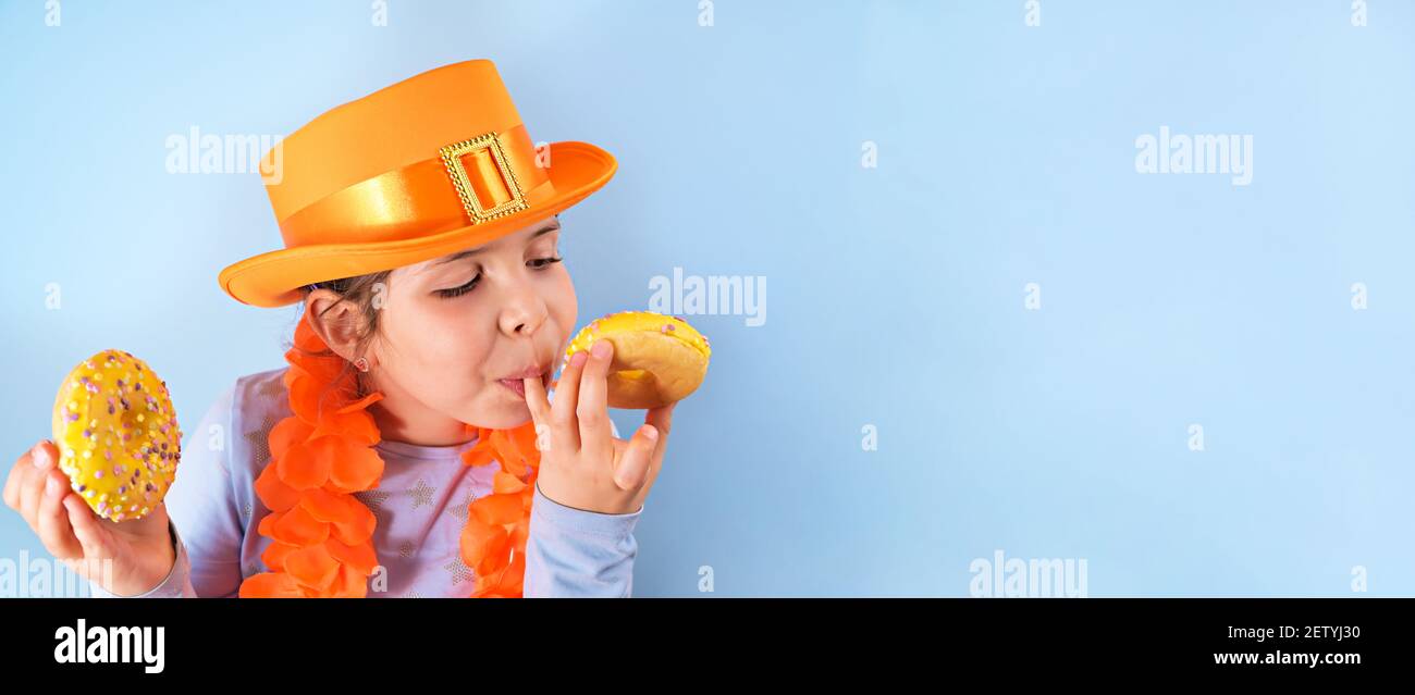 King's Day in Olanda. Festa tradizionale il 27 aprile nei Paesi Bassi. Una bambina in un cappello arancione festivo su uno sfondo blu mangia ciambelle colorate. Banner, formato lungo. Spazio di copia Foto Stock