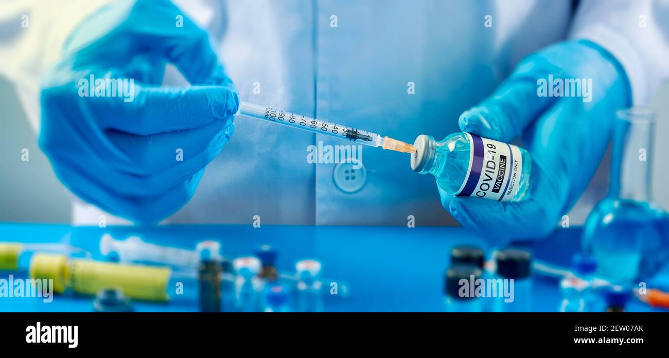 chiusura di un operatore sanitario o di laboratorio, indossando guanti blu, riempiendo una siringa da un flaconcino simulato di vaccino covid-19, su un tavolo Foto Stock
