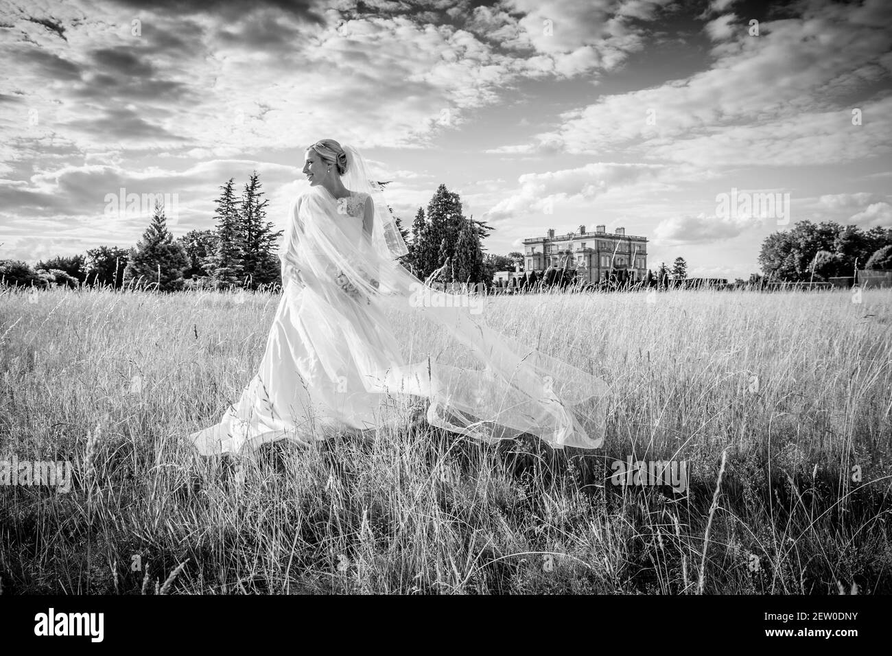 Sposa in piedi nel mezzo di un campo d'erba con una brezza che soffia il suo velo intorno. È sotto un grande cielo con un palazzo georgiano sullo sfondo Foto Stock