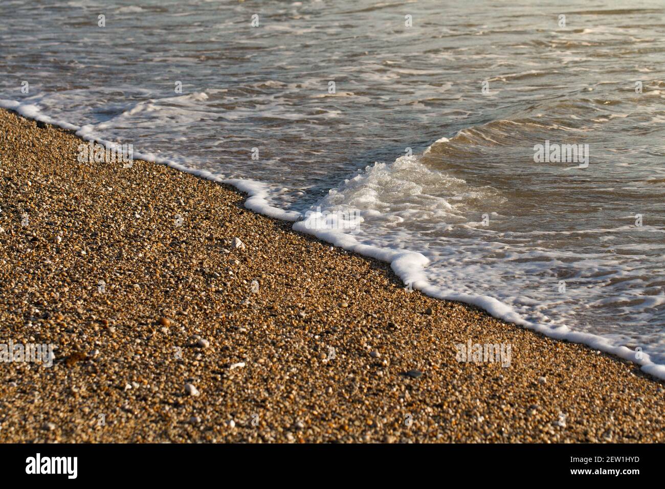 Primo piano di una spiaggia di ciottoli illuminata da una dorata risplendere con onde delicate che lambano il bordo dell'acqua un tranquillo sfondo sul mare Foto Stock