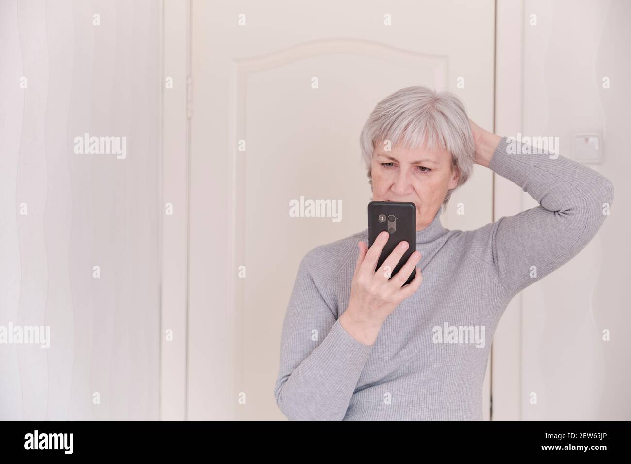 Una donna anziana con capelli grigi in un dolcevita grigio che guarda attentamente lo smartphone a casa. Il momento dello stile di vita. Foto Stock