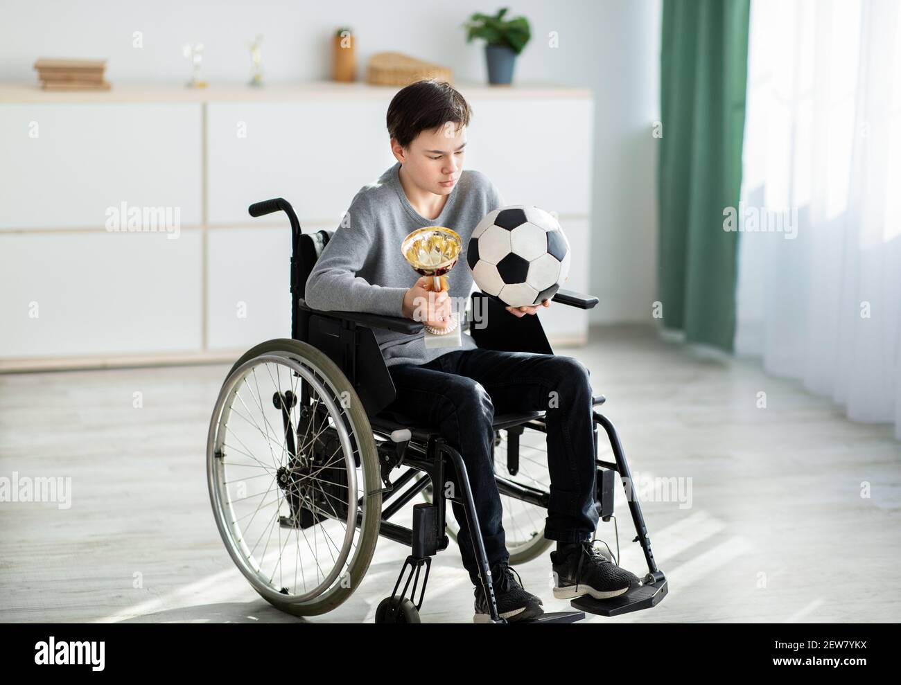 Teen giocatore di calcio handicappato con palla e trofeo seduto in sedia a rotelle, sentendosi depresso per la sua ferita a casa Foto Stock