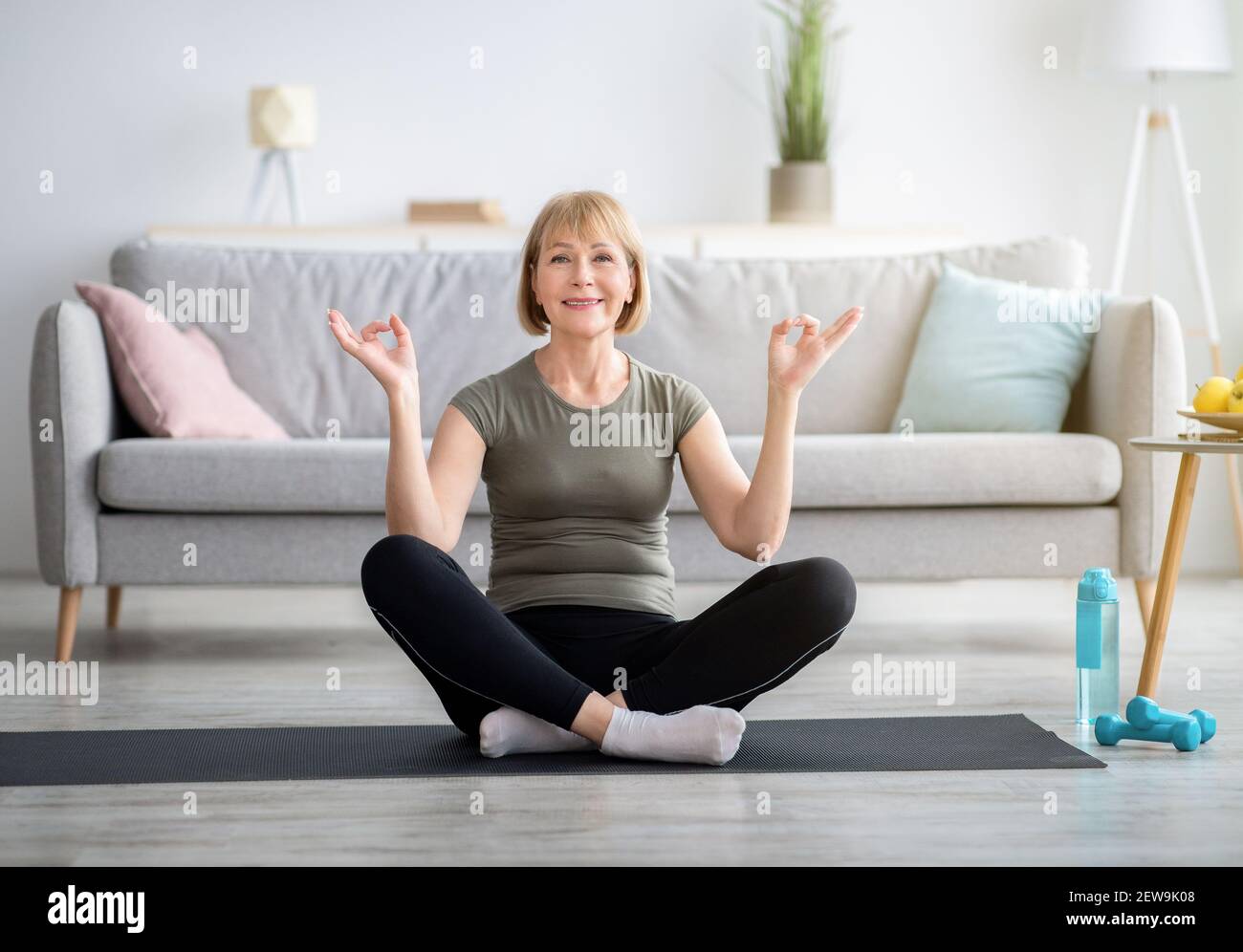 Donna anziana equilibrata seduta in posa lotus e facendo mudra gyan con le mani, meditando sul tappeto sportivo a casa Foto Stock
