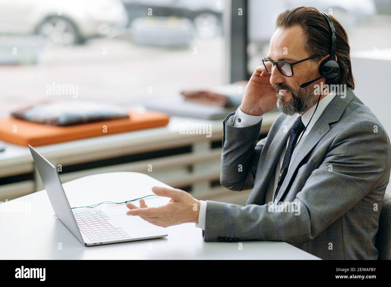 Bell'uomo d'affari anziano in cuffia sta usando il laptop, avendo una videoconferenza. Felice manager maschile che parla con i colleghi, sorridendo, discutendo di progetto aziendale Foto Stock