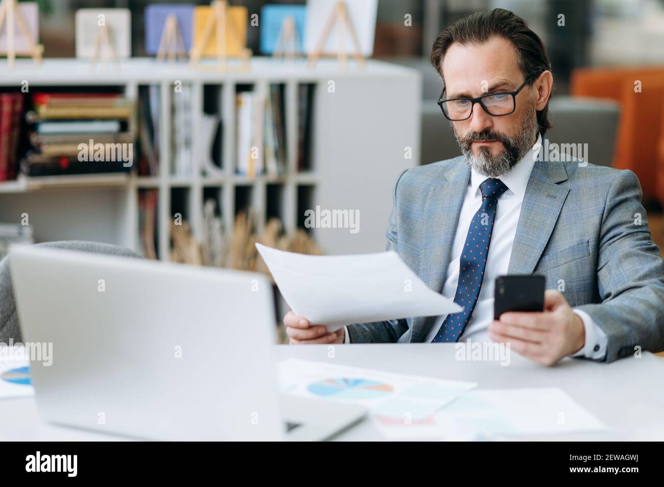 Un uomo d'affari concentrato e con portatore negli occhiali utilizza lo smartphone, chiacchierando con i colleghi. Uomo d'affari serio seduto alla scrivania, lavorando su progetto importnat Foto Stock