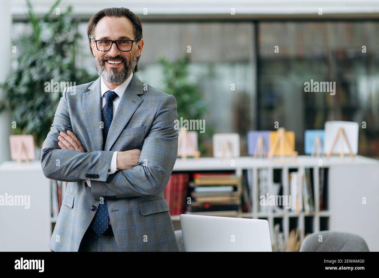 Ritratto di un convinto imprenditore di mezza età in abito elegante e occhiali in piedi in ufficio moderno, guardando la fotocamera e sorridente amichevole Foto Stock