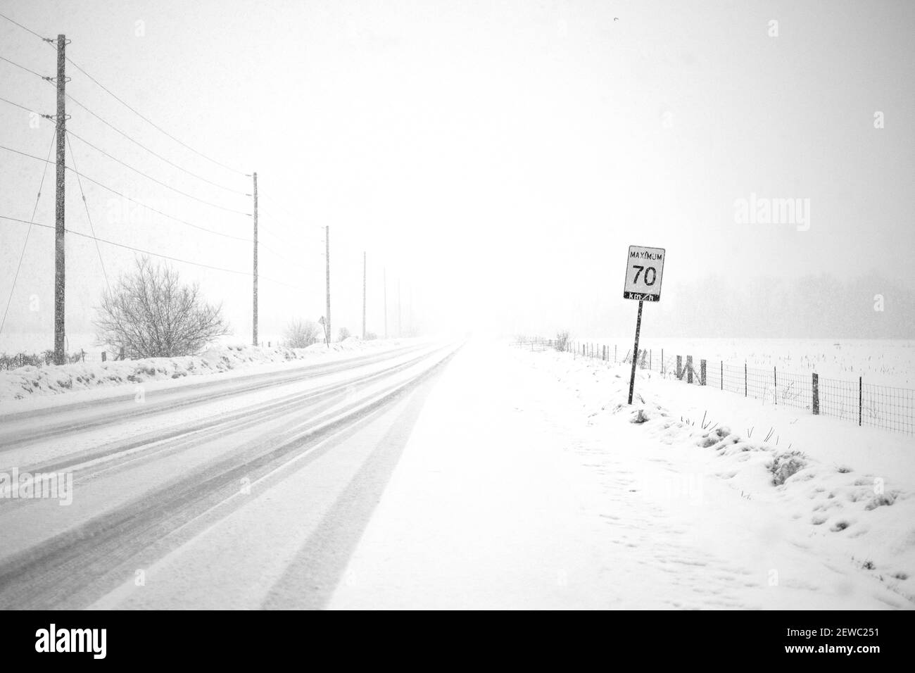 Durante le condizioni di whitout di una bizzard, le linee elettriche lungo una strada rurale svaniscono nella neve. Un segnale del limite di velocità indica un limite di 70 km/h. Foto Stock