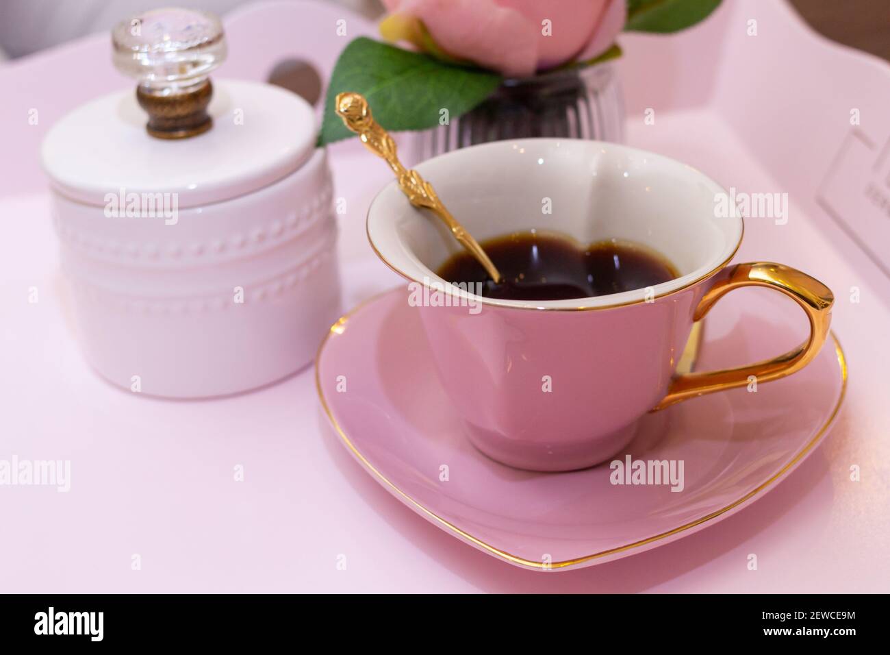 Una tazza di caffè rosata a forma di cuore in uno stile aristocratico con un cucchiaio dorato in esso con una ciotola di zucchero e. fiori sullo sfondo Foto Stock
