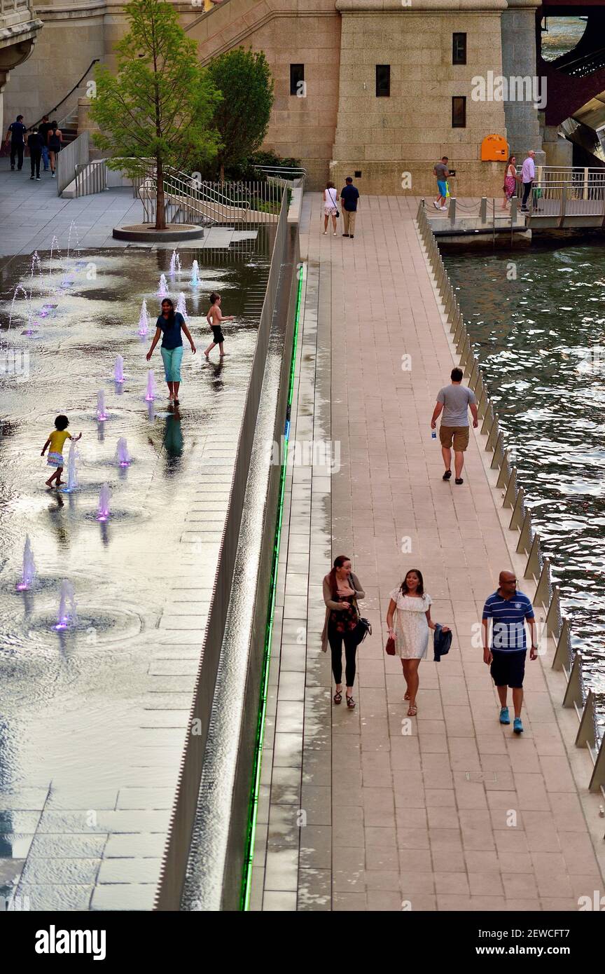 La gente si sposta tranquillamente lungo il Chicago Riverwalk mentre bambini e genitori girovagano nella fontana attiva lungo la passerella. Foto Stock