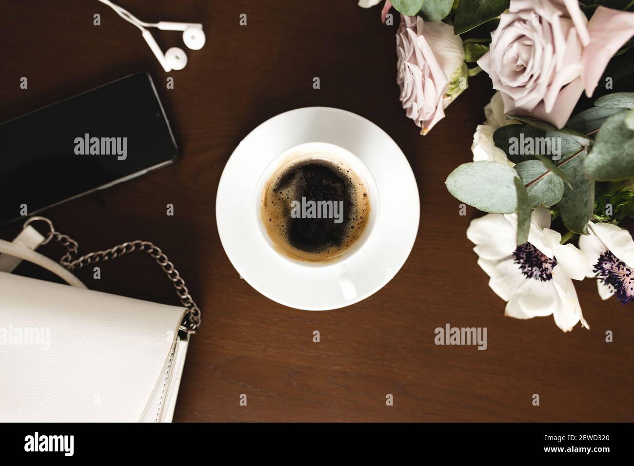 Una tazza di caffè con un bouquet di tenera anemone, rami di eucalipto e rose viola pastello, una borsa donna, cuffie e un telefono su un legno marrone Foto Stock