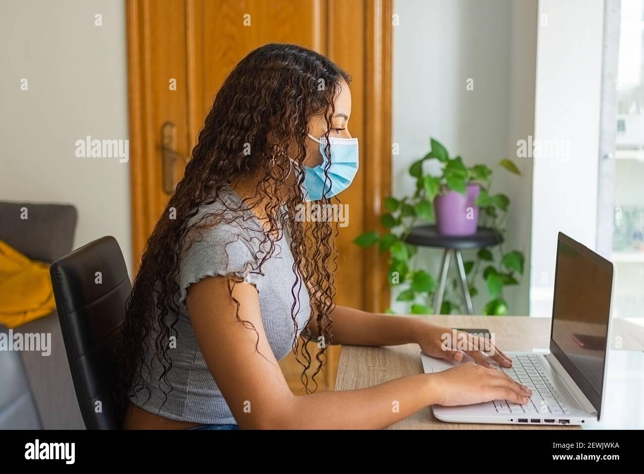 ragazza con una maschera sul viso utilizzando il computer sul tavolo del soggiorno mentre sta telelavoro da casa Foto Stock