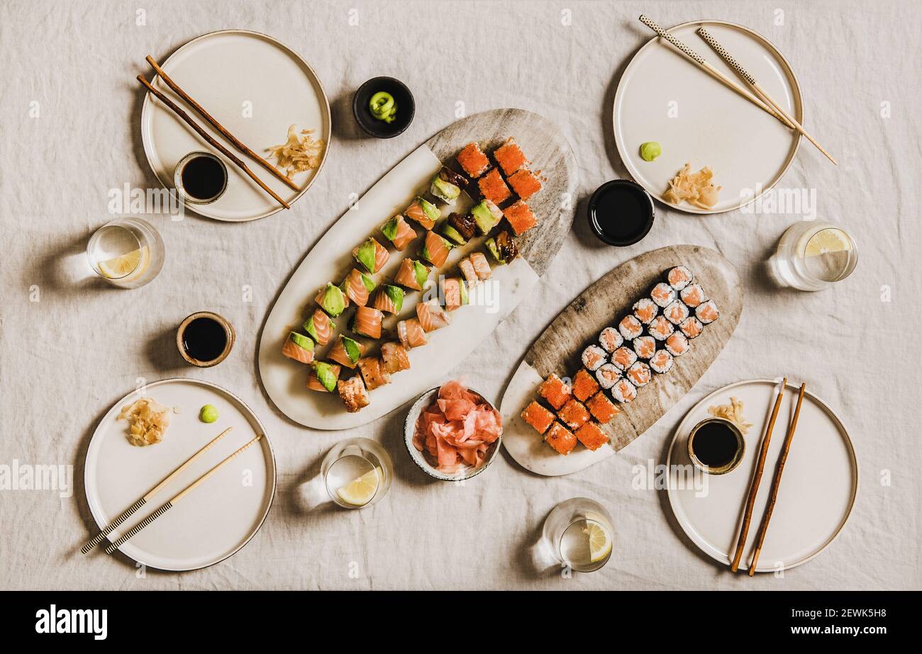 Festa con cena casalinga di Lockdown con sushi Giapponese dal servizio di consegna o da asporto. Piatto di salmoni, granchi, gamberi, involtini vegani, wasabi, zenzero ana limone acqua su sfondo chiaro, vista dall'alto Foto Stock