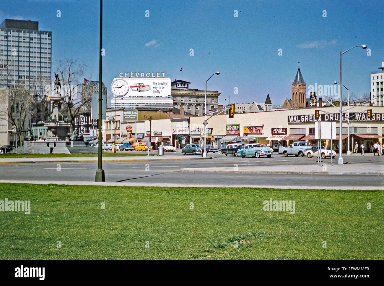 Downtown Denver, Colorado, USA c. 1960. Questa vista dal Civic Central Park è l'incrocio tra West Colfax Avenue e North Broadway. A sinistra si trova la Fontana dei pionieri (o Monumento dei pionieri), una fontana e scultura di Frederick William MacMonnies. La statua è di Kit Carson. Le auto passano accanto ai negozi e alle aziende di questo incrocio. Un cartellone gigante Chevrolet sorge sopra di loro. Dice che è appena dopo le 15. L'edificio centrale YMCA è visibile sullo sfondo. Oggi l'YMCA è oscurata dall'One Civic Center Plaza, un alto edificio a 22 piani costruito dove un tempo sorgevano i negozi. Foto Stock