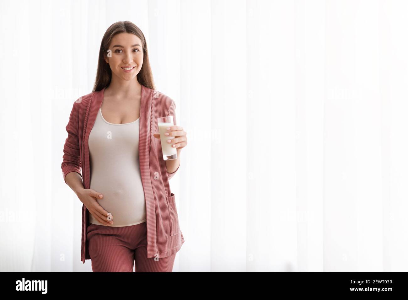 Bella donna incinta con bicchiere di latte in posa vicino alla finestra A casa Foto Stock