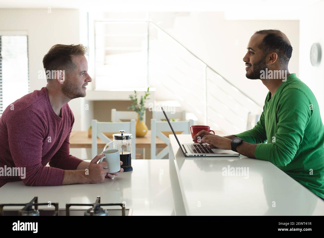 Multi etnica gay maschio coppia sorridente e seduto in cucina bere caffè e utilizzare il computer portatile a casa Foto Stock