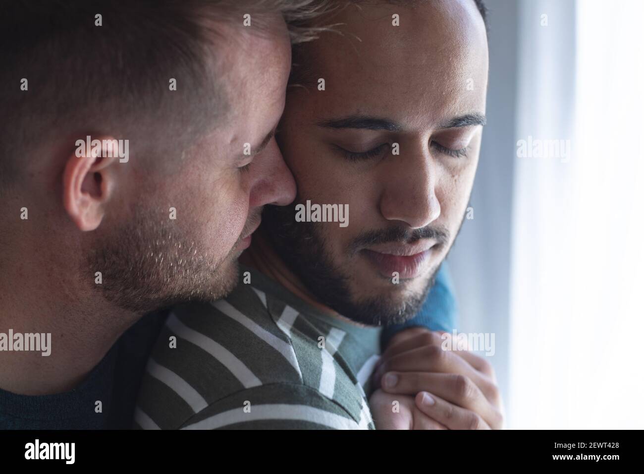 Multi etnica gay maschio coppia in piedi con gli occhi chiuso hugging a casa Foto Stock