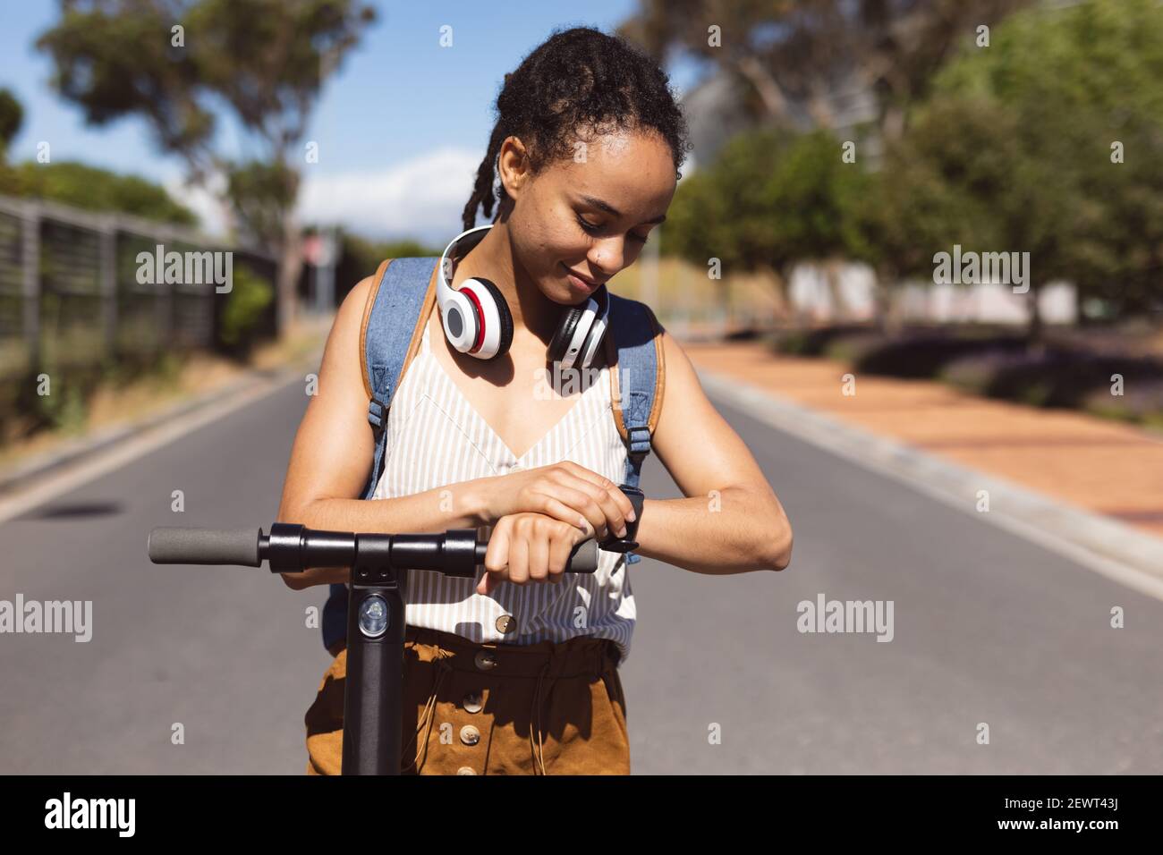 Donna afroamericana sorridente che indossa le cuffie in piedi su scooter usando smartwatch in strada Foto Stock