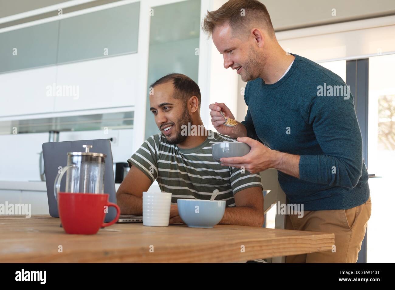 Multi etnica gay maschio coppia sorridente e mangiare la colazione e. utilizzo di un computer portatile a casa Foto Stock