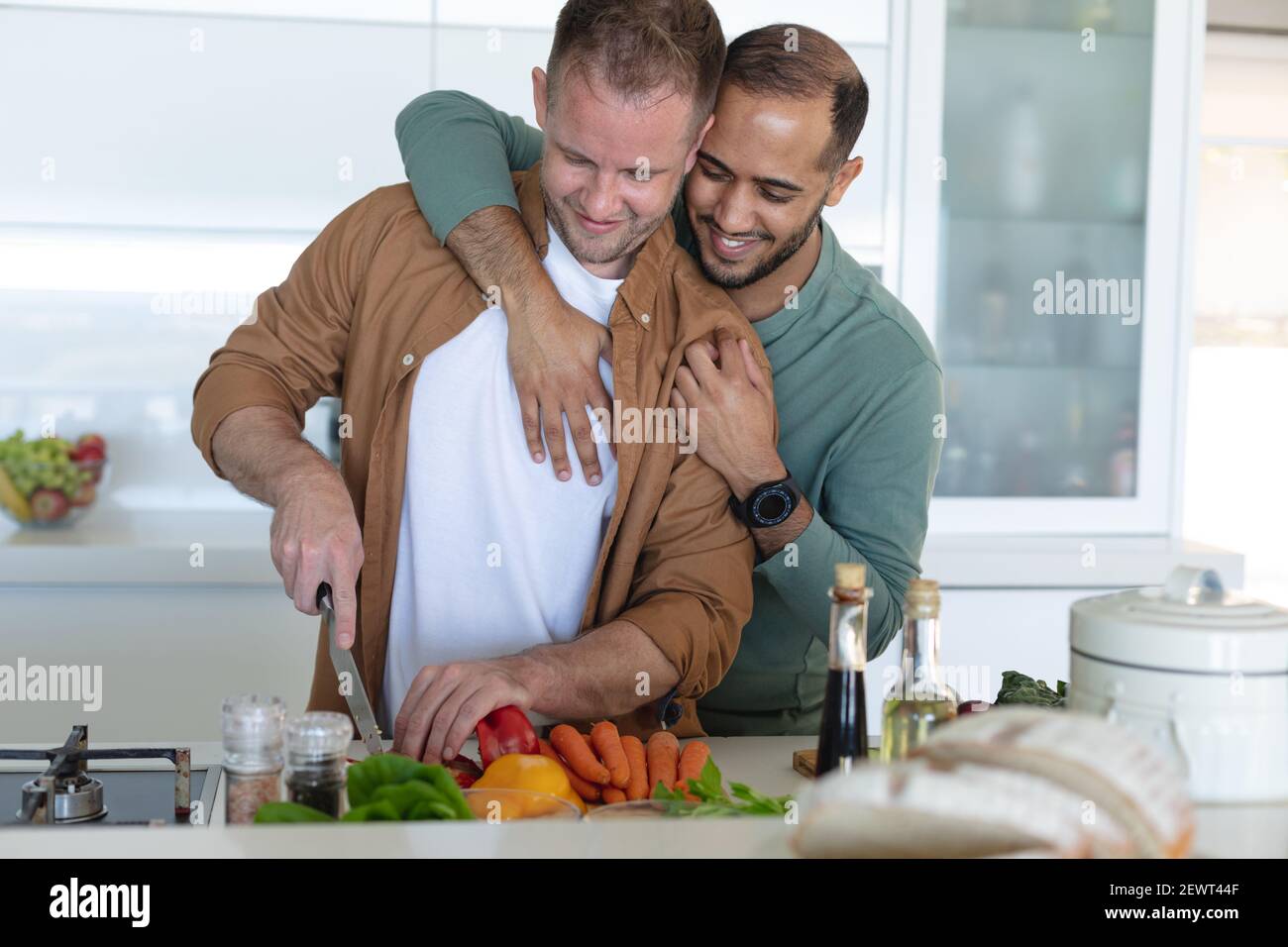 Multi etnica gay maschio coppia sorridente, preparando cibo e abbracciando a casa Foto Stock