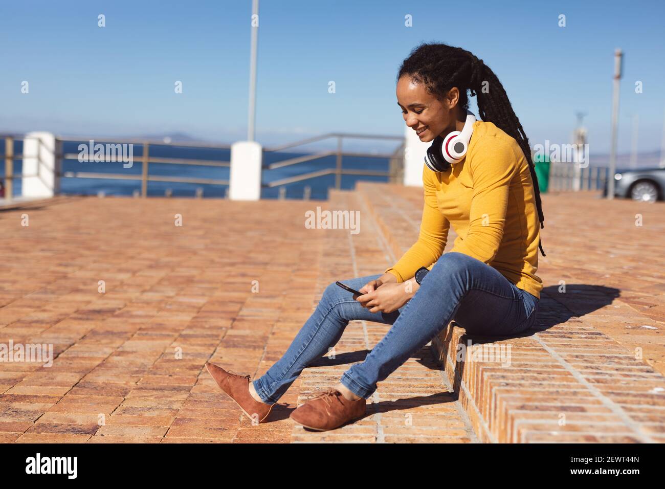 Donna afroamericana sorridente, indossando le cuffie seduti con smartphone sulla passeggiata sul mare Foto Stock