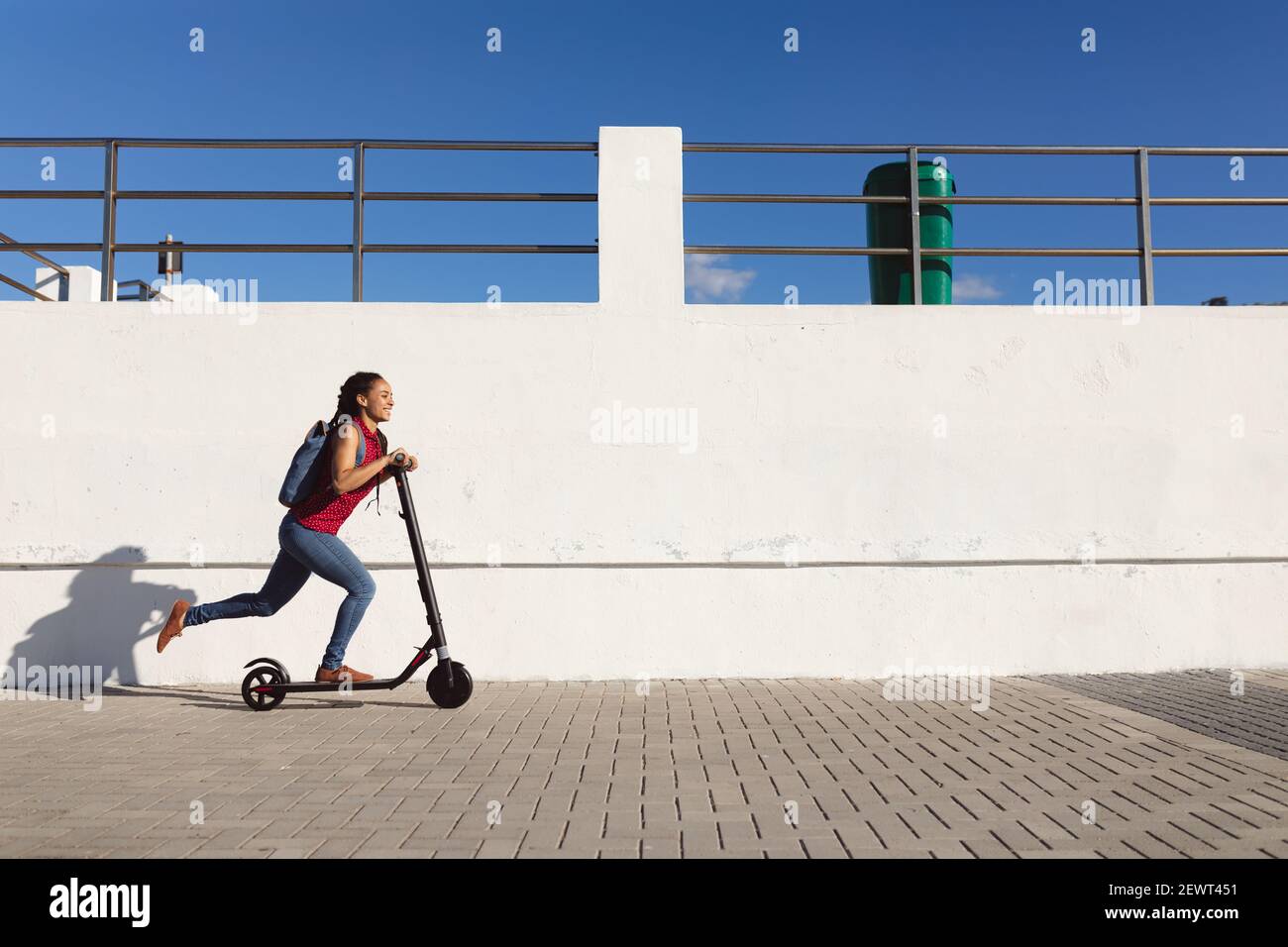 Donna afroamericana sorridente che indossa uno zaino in sella a uno scooter sulla passeggiata vicino al mare Foto Stock