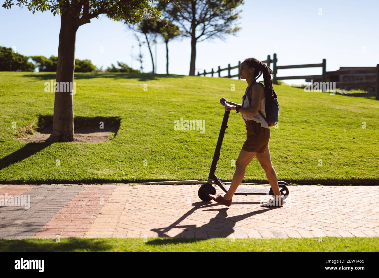 Felice donna afroamericana che indossa zaino che cammina con uno scooter nel parco Foto Stock