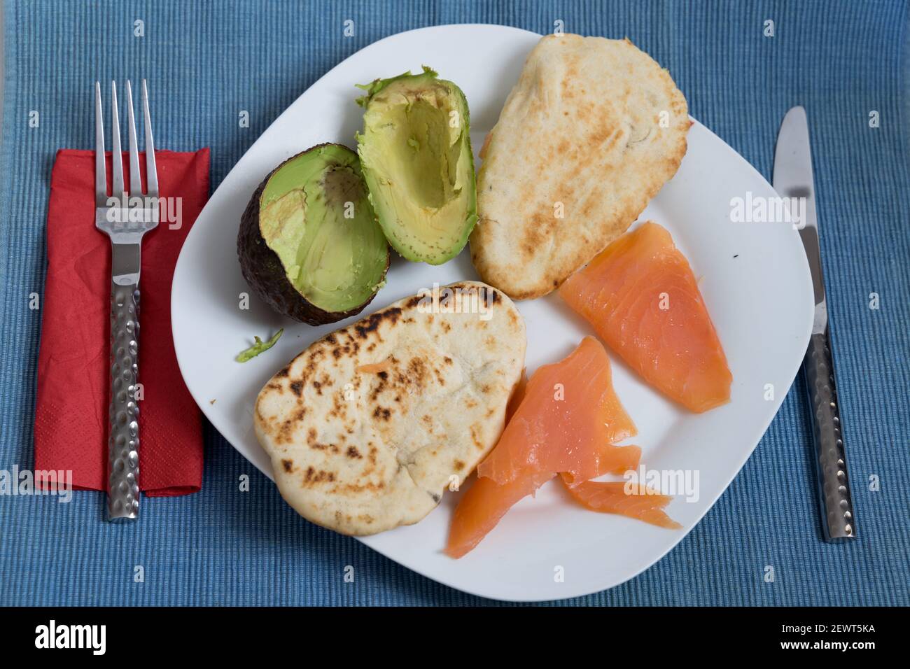 due pani naan tostati con salmone e mezzo avocado su un piatto bianco , tovagliolo rosso, forchetta e coltello , su tappetino blu Foto Stock
