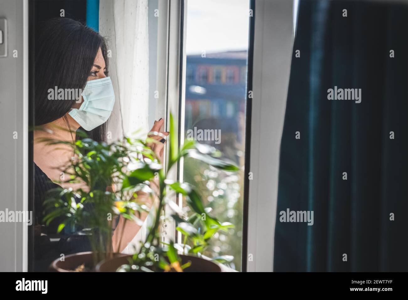 Giovane donna che guarda fuori la finestra con maschera medica a casa. Covid-19 e nuovo concetto normale. Foto Stock