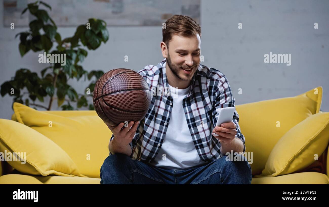 felice tifoso di basket che tiene la palla e guarda la partita sullo smartphone Foto Stock