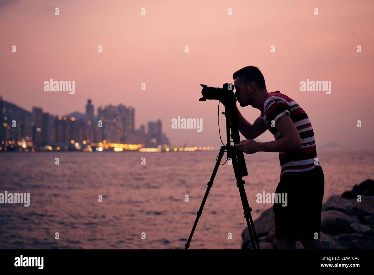 Silhouette di fotografo con treppiede. Giovane uomo che fotografa lo skyline urbano. Hong Kong al tramonto. Foto Stock