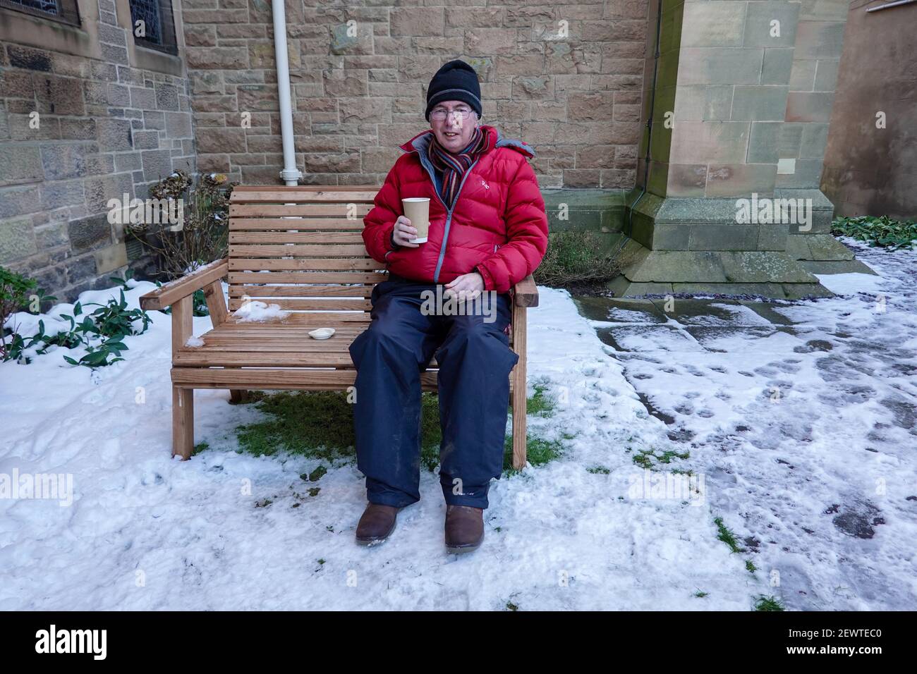 Uomo seduto sulla panca che beve caffè. Costretto a sedersi all'esterno, nella neve e nel ghiaccio, durante il blocco a causa della pandemia del coronavirus Foto Stock