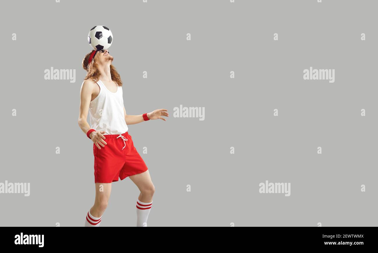 Giovane uomo in abbigliamento sportivo equilibrando palla di calcio sulla fronte isolato su sfondo grigio Foto Stock