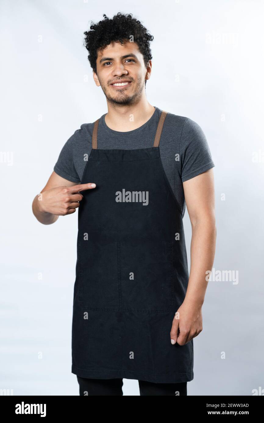 Grembiule nero mockup - giovane ispanico che punta al grembiule sorridente su sfondo bianco - giovane lavoratore - modello per testo sui vestiti Foto Stock