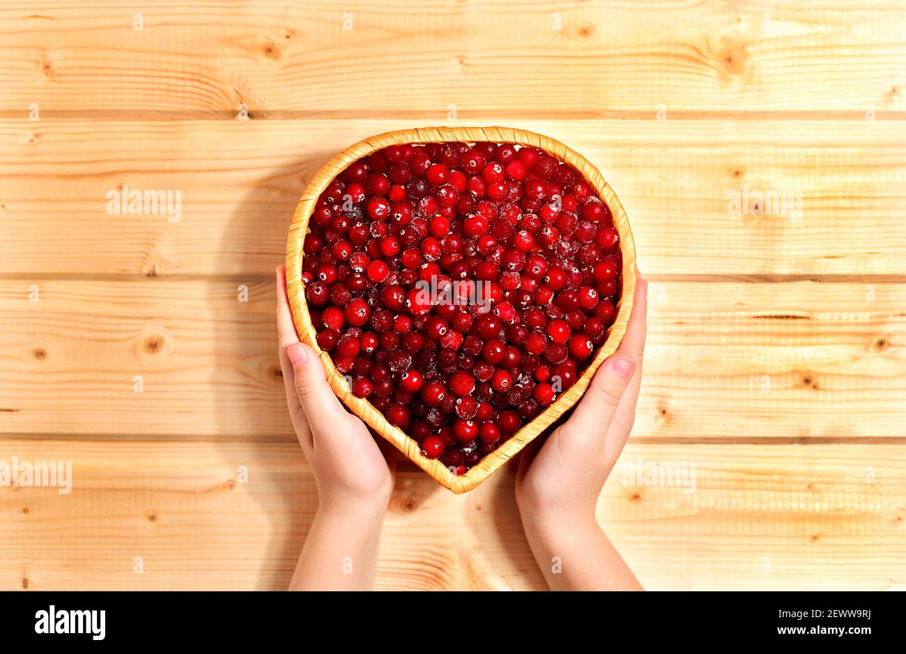 Bacche di mirtillo rosso surgelate, in un vimini di canestro di vite a forma di cuore. Con cura in mano su uno sfondo caldo di legno. Foto Stock